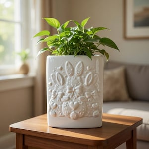 Peut inclure: Un pot de fleurs rectangulaire en céramique blanche avec des motifs de lapins et de cœurs en relief, rempli de plantes vertes à feuilles. Le pot est posé sur une table en bois, avec une fenêtre et un canapé en arrière-plan. Le pot mesure environ 20 cm de haut.