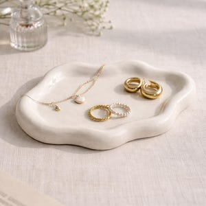 May include: A white, organically shaped ceramic tray holds gold jewelry. A delicate gold necklace with a heart pendant, two gold hoop earrings, and two rings are displayed. A small perfume bottle and flowers are in the background.