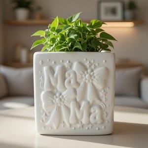 May include: A square white ceramic planter with the word "Mama" embossed on the front, decorated with bows and stars. The planter is filled with vibrant green leafy plants. The planter is sitting on a light-colored surface.