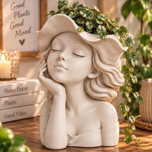 Peut inclure: Un pot de fleurs en résine couleur crème en forme de tête et d'épaules, portant un chapeau à larges bords. La figure a les yeux fermés et une main posée contre la joue. Une plante verte retombante aux petites feuilles en cœur pousse du chapeau.