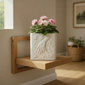 Puede incluir: Una maceta cuadrada de cerámica blanca con un diseño en relieve de una mano sosteniendo otra mano, llena de plantas con flores rosas y blancas. La maceta está sobre una estantería flotante de madera clara.