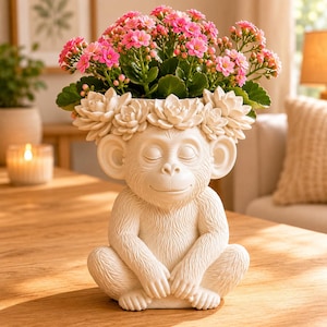 May include: A cream-colored monkey-shaped planter with a serene expression, adorned with a floral crown, holds a vibrant arrangement of pink flowers and green foliage. The monkey is seated with its hands clasped, creating a whimsical and decorative piece.