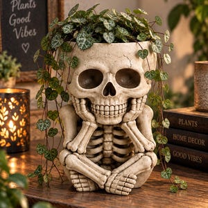 May include: A beige skull-shaped planter featuring a skeleton figure with hands resting on its chin. The planter is filled with a trailing plant with small heart-shaped green leaves. The skull has a textured, weathered finish with deep eye sockets.