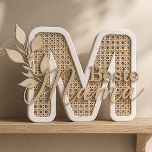 May include: A decorative wooden letter "M" with a white frame and woven cane center. The words "Beste Mama" are in a light brown script across the front. A sprig of leaves is attached to the left side. The item is displayed on a wooden shelf.