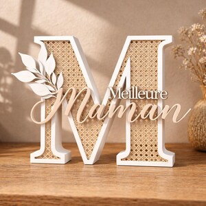 Puede incluir: Una letra "M" decorativa de madera blanca con un centro de ratán tejido. La palabra "Meilleure Maman" está escrita en cursiva en la parte delantera. Un diseño de hojas blancas está adherido al lado izquierdo de la letra.