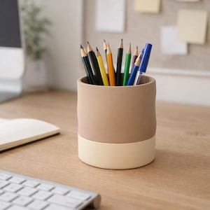 Peut inclure: Un pot à crayons cylindrique beige et crème rempli de crayons et de stylos de couleurs variées. Le pot est posé sur un bureau en bois, avec un carnet et un clavier en arrière-plan.