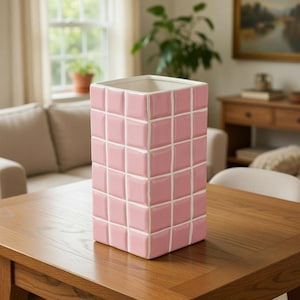 May include: A pink ceramic vase with a square shape and a grid pattern. The vase is made of individual squares with white grout lines. The interior of the vase is white. The vase is sitting on a wooden table.