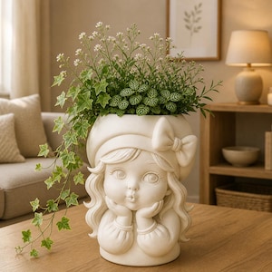 May include: A cream-colored ceramic planter shaped like a girl's face, with a bow on her hat and hands supporting her chin. The planter is filled with green plants and white flowers, creating a decorative indoor accent.