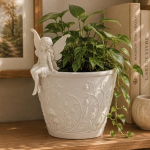 May include: A white ceramic planter with a fairy figurine, filled with green leafy plants. The planter features embossed floral and mushroom designs, with a crescent moon detail. The fairy is seated on the planter's edge.