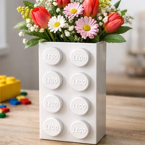 Peut inclure: Un vase en céramique blanc en forme de brique LEGO, rempli d'un bouquet de tulipes rouges, de marguerites roses et blanches et de fleurs jaunes. Le mot "LEGO" est en relief sur le vase.