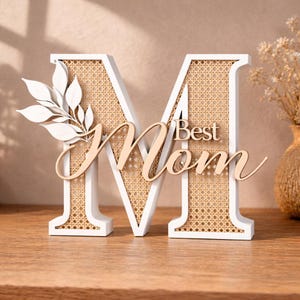 Op de afbeelding: Een decoratieve witte houten letter "M" met een geweven rotan midden en de woorden "Best Mom" in een cursief lettertype. Een wit bladerontwerp is aan de linkerkant bevestigd. Een cadeau voor Moederdag.