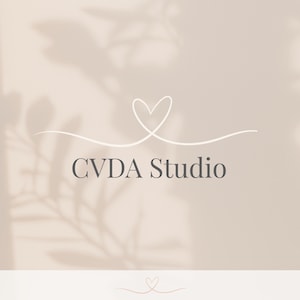 May include: A logo design featuring the text "CVDA Studio" in a dark gray font, with a white heart-shaped design above the text. The background is a light beige color with subtle shadow details.