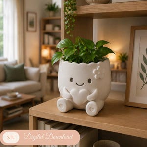 Op de afbeelding: Een witte keramische plantenpot in de vorm van een stripfiguur, met een hart en een lachend gezicht. De pot bevat groene bladplanten en staat op een houten plank. De tekst "Digital Download" is zichtbaar.