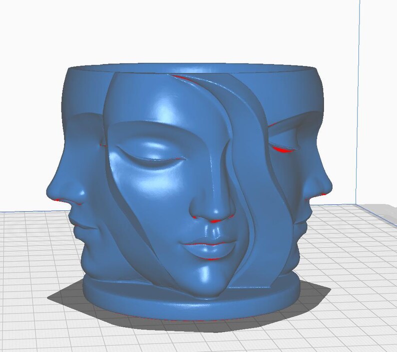 May include: A blue cylindrical planter featuring the sculpted faces of three women. The faces are in profile and have closed eyes and serene expressions. The planter is a solid blue color and sits on a small base.