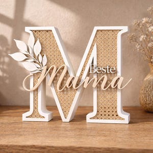 May include: A white wooden letter 'M' with a woven rattan center and the words 'Mama' and 'Beste' in cursive script. A decorative leaf design is attached to the left side. The piece is a decorative item.