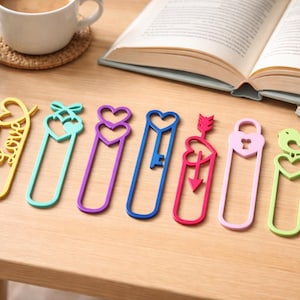 Pode incluir: Uma coleção de marcadores coloridos com vários designs, incluindo corações, chaves e pássaros. Um marcador tem a palavra "love" em uma fonte cursiva. Os marcadores estão dispostos em uma superfície de madeira perto de uma xícara de café e um livro aberto.
