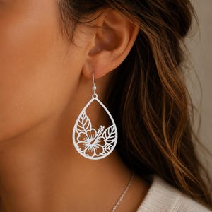May include: Silver-toned teardrop earrings with a cut-out hibiscus flower and leaf design. The earrings have a hook closure and are worn on the ear. The floral design is intricate and detailed, showcasing a tropical theme.