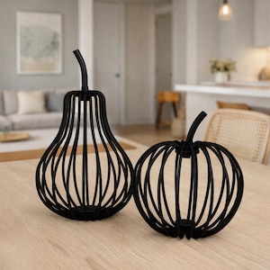 May include: Two black metal decorative sculptures, one pear-shaped and the other pumpkin-shaped, sit on a light wood surface. The sculptures are made of vertical metal rods, creating an open, cage-like design. The background shows a living room setting.