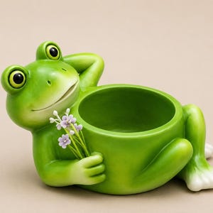 Peut inclure: Un cache-pot en forme de grenouille vert vif représentant une grenouille allongée, une main derrière la tête et l'autre tenant un petit bouquet de fleurs violettes. L'ouverture circulaire pour une plante est intégrée au corps.