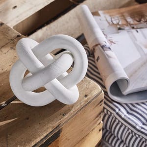May include: A white knot sculpture, crafted from a solid material, sits on a wooden surface. A rolled-up magazine and striped fabric are in the background. The decorative object has a simple, modern design, suitable for home decor.