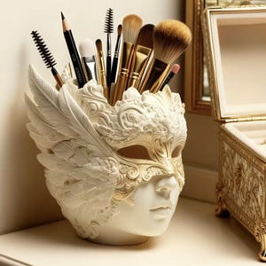 May include: A white and gold decorative mask-shaped makeup brush holder. The holder features a face design with intricate detailing, including wings and a mask. The holder is filled with various makeup brushes and pencils.
