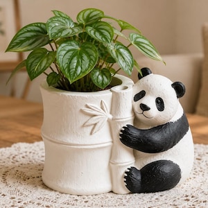 May include: A white ceramic panda planter holding a bamboo stalk, with a green leafy plant inside. The panda has black ears, eyes, and limbs. The planter is on a lace doily.