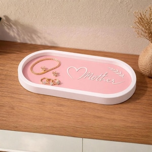 Peut inclure: Un plateau à bijoux ovale blanc et rose avec le mot "Mother" en écriture cursive. Le plateau contient un bracelet, des boucles d'oreilles et des bagues dorés. Posé sur une surface en bois.