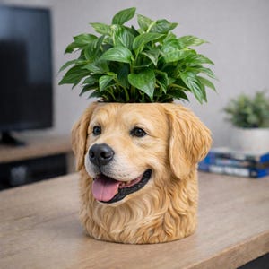 Peut inclure: Un pot de fleurs en forme de tête de chien de couleur beige, avec une expression joyeuse, la langue sortie, et une plante verte feuillue qui pousse du haut. Le pot est posé sur une table en bois.