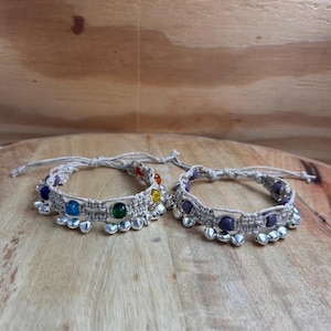 May include: Two macrame bracelets with colorful beads and silver-toned accents. One bracelet features a rainbow of beads, while the other has purple beads. The bracelets are displayed on a wooden surface.