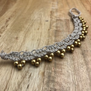 May include: A beige macrame bracelet with gold beads. The bracelet is made with a knotting technique and has a tassel at the end.