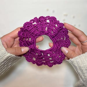 Puede incluir: Un coletero de ganchillo en color magenta vibrante con un diseño circular de volantes y un intrincado punto calado en toda la tela. El accesorio tiene un centro elástico fruncido y un borde exterior decorativo festoneado.