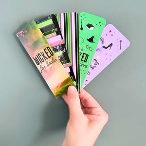 May include: A hand holding a fan of colorful bookmarks. The bookmarks feature various designs, including text that reads "Wicked for books", a witch hat, a high heel, and a magic wand. The color palette includes green, pink, and purple.