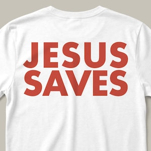 Puede incluir: Camiseta blanca con el texto rojo "JESUS SAVES" impreso en la parte delantera. El texto está en negrita, con una fuente sans-serif. La camiseta es probablemente de algodón.