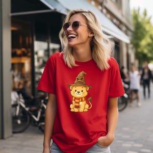 May include: A red t-shirt featuring a cartoon lion wearing a brown wizard hat and a striped scarf. The lion is sitting, with a tail and a smiling face. The shirt is made of a soft material.