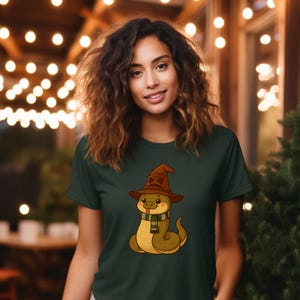 May include: Dark green t-shirt featuring a cartoon snake wearing a brown witch hat and a green and white striped scarf. The snake is sitting down with a friendly expression. The shirt is made of a soft material.