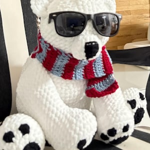 Può includere: Un peluche fatto a mano di orso polare bianco con occhiali da sole neri e una sciarpa a righe rosse, bianche e blu. L'orso ha cuscinetti neri e si trova su una superficie a strisce bianche e nere.