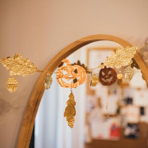 May include: A gold-colored Halloween garland featuring a carved pumpkin, oak leaves, acorns, and a dangling leaf. The garland is strung across a wooden-framed mirror, creating a festive autumn decoration.