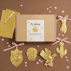 Puede incluir: Un kit de estampado de lámina DIY con una caja marrón y detalles de lámina dorada. El kit incluye piezas decorativas de color dorado, como una mano, un cactus, una calavera y un pájaro. La caja tiene una etiqueta que dice "Pr Aline Creative Kits & Decor DIY Foil Embossing Kit."
