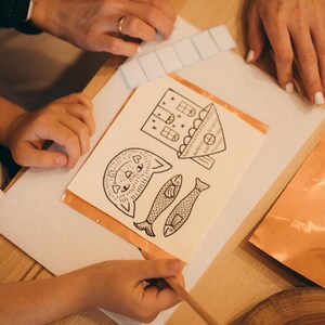 May include: A collection of illustrated paper cutouts featuring a house, a cat, and two fish. The drawings are in black ink on a cream-colored background. The paper is placed on a wooden surface, with hands and a pencil visible.