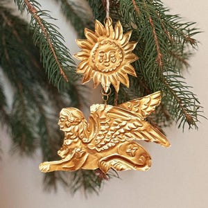 May include: Gold-colored Christmas ornaments. One is a sun with a smiling face, and the other is a winged sphinx. The ornaments are hanging from a pine branch. The ornaments are made of metal and have intricate details.