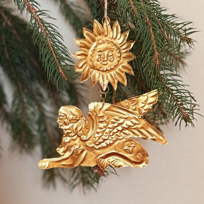 May include: Gold-colored Christmas ornaments. One is a sun with a smiling face, and the other is a winged sphinx. The ornaments are hanging from a pine branch. The ornaments are made of metal and have intricate details.