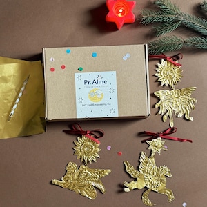 Puede incluir: Una caja de kit de manualidades con el texto "Pr. Aline Creative Kits & Decor" y "DIY Foil Embossing Kit". Adornos metálicos dorados con forma de sol, estrella y criatura alada con cintas rojas. También hay una vela roja en forma de estrella encendida y una ramita de hoja perenne.