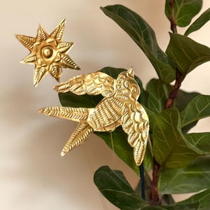 Può includere: Due oggetti decorativi color oro: una stella e un uccello in volo. La stella ha raggi appuntiti e un cerchio centrale. L'uccello ha piume di ali e coda dettagliate. Gli oggetti sono posti su uno sfondo di foglie verdi.
