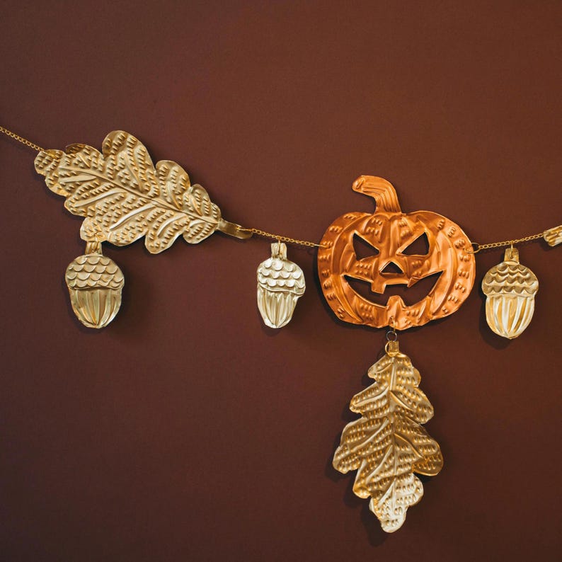 May include: A decorative fall garland featuring a carved pumpkin, acorns, and leaves in gold and copper tones. The garland is strung on a delicate gold chain, perfect for autumn decor.