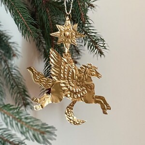 May include: A gold-colored Pegasus ornament with detailed wings and a crescent moon shape. The ornament is suspended from a star-shaped top, hanging from a white string, and set against a backdrop of green pine needles.
