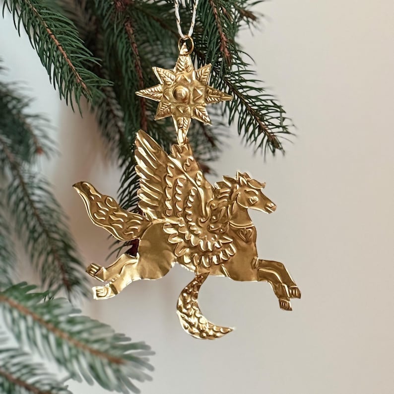 May include: A gold-colored Pegasus ornament with detailed wings and a crescent moon shape. The ornament is suspended from a star-shaped top, hanging from a white string, and set against a backdrop of green pine needles.