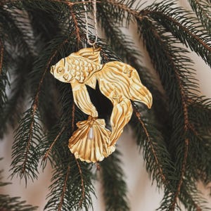 May include: A gold-colored fish ornament with a seashell and pearl detail, hanging from a white string. The fish has detailed scales and fins, and is suspended against a backdrop of green pine needles.