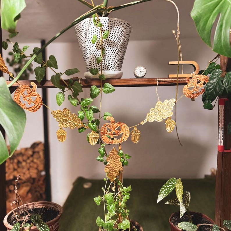 May include: A festive autumn garland featuring carved pumpkins, oak leaves, and acorns in shades of gold and brown. The garland is strung on twine and is displayed among potted plants, creating a seasonal decorative display.