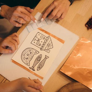 May include: A craft project in progress, featuring a drawing of a cat, house, and fish. The drawing is on a white paper, with copper tape around the edges. Clear glass pieces are also present, along with copper sheets.
