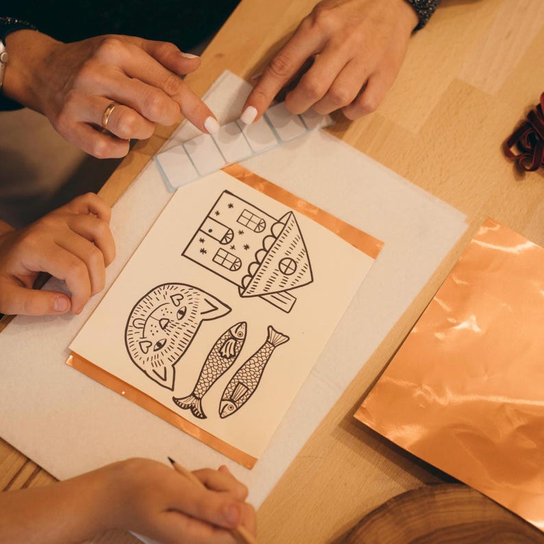 May include: A craft project in progress, featuring a drawing of a cat, house, and fish. The drawing is on a white paper, with copper tape around the edges. Clear glass pieces are also present, along with copper sheets.